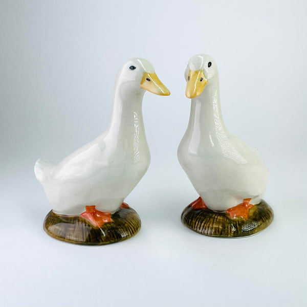 Two tall smooth white ducks standing separately on their own bases which look like a round piece of earth. They both have orange feet, set firmly on the ground with their little tummies resting on them. Yellow beaks, with black nostrils and black eyes. They are standing looking forward.