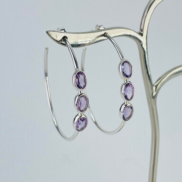 Large silver hoop earrings with three oval faceted amethyst stones set into the front of the hoop. The stones are wider than the hoop and set into a frame of silver.
