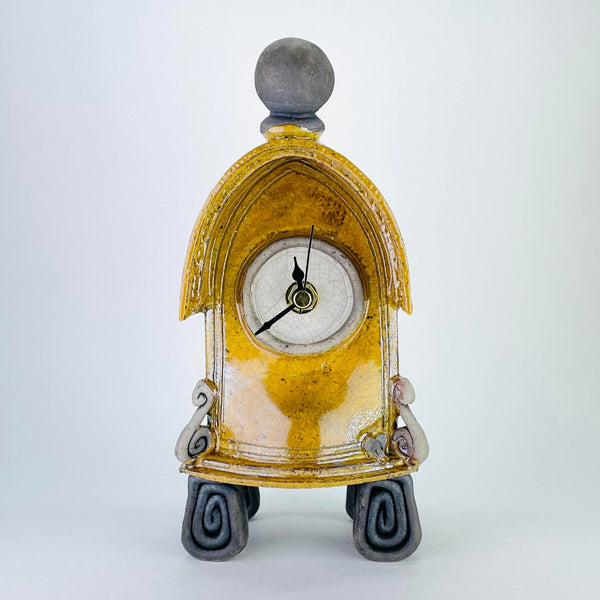 This quirky clock has the shape of a gothic arch or the porch of an old church, with a little roof effect on the top. It has four feet formed of square spirals of matte dark grey, a scroll at each side of the front, with a tiny mouse head peering out! It is topped with a matte grey orb. The main body is a shiny ochre colour, the central round face is white and it has a minute, an hour and a second hand - all black.