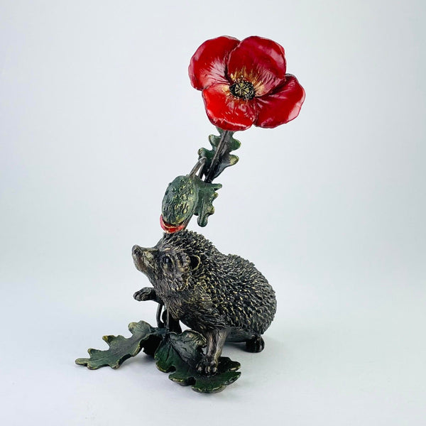 A little hedgehog is standing on a leaf at the bottom of a poppy plant. The stem has two more pairs of leaves set opposite each other with a flower in bud towards the top. The bud is red. At the top is a flower in full bloom, with four big red petals and yellow and black highlighting inside. The hedgehog is well detailded, standing slightly up with its nose in the air and a cute face,