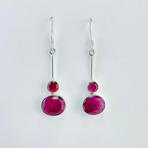 Dark pink, faceted ruby quartz stones. There are two stones on each earring. On the bottom of a silver stalk is a round ruby quartz stone, beneath this is a larger oval shaped stone, sitting horizontally rather than vertically. The silver stalk is attached to the silver hook by a plain silver ring which is part of the stalk.