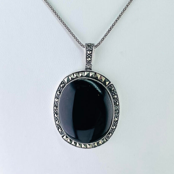 A shiny black oval onyx stone is surrounded by little marcasite stones cut in two shapes. The stones at the top and bottom are square cut, the ones on the side are circular. The pendant is attached to a marcasite set bail and is hanging off an oxidized chain.