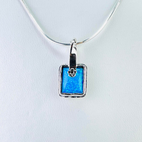 A bright sparkly blue opal stone is set within a double silver frame with a slightly wavy edge. At the top of the square is a very small five petalled silver flower which looks like it is holding the silver bail in place . The bail is a shiny fairly wide band of silver which twists at the top where the chain goes through.
