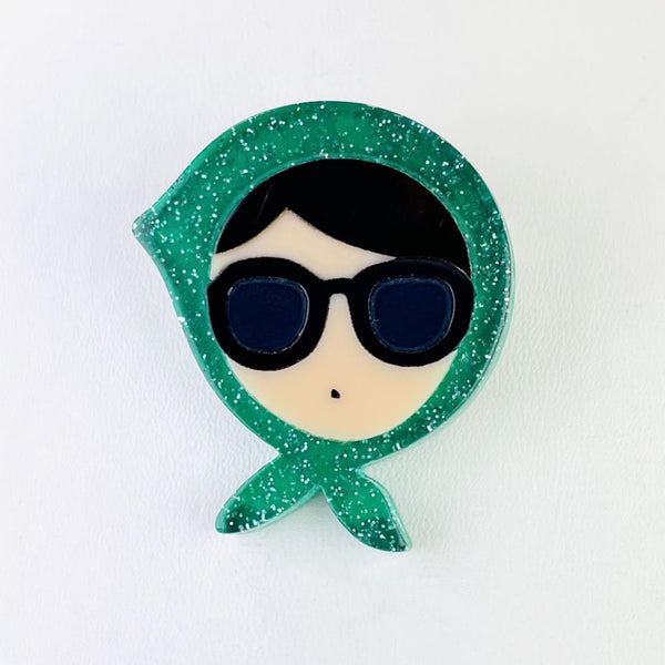 A cream female face forms the centre of this brooch. She has dark hair showing as a fringe with a side parting, is wearing large sunglasses and just has a dot as a mouth.  She has a green glittery scarf tied around her head knotted under her chin, a little like the Queen would wear.