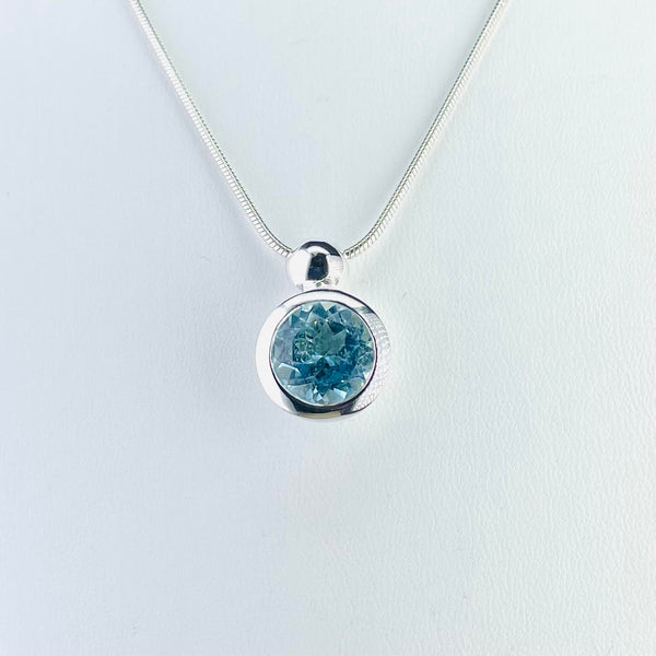 A very simple round pendant on a silver chain. The round pale, but bright, blue faceted stone is simply framed in shiny silver. It is topped by a round silver bail which the chain is threaded through.