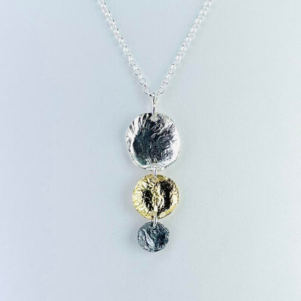 Three discs decreasing in size as they go down are connected by small silver rings. All have a textured finish. The top is shiny silver, the middle is gold plated and shiny and the bottom in dark oxidizes silver. Attached to a fine silver link chain by a delicate plain bail.
