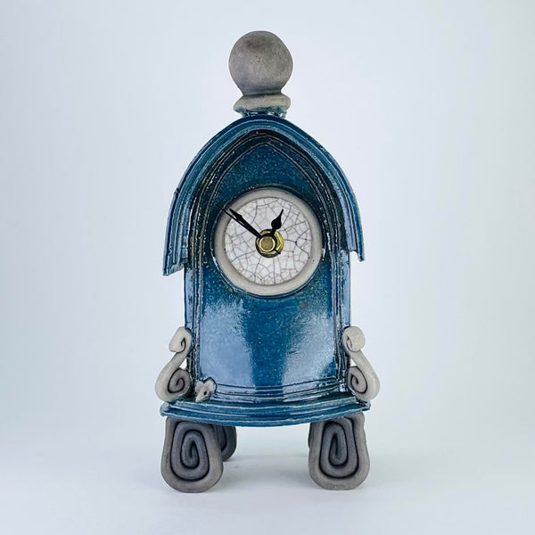 This quirky clock has the shape of a gothic arch or the porch of an old church, with a little roof effect on the top. It has four feet formed of square spirals of matte dark grey, a scroll at each side of the front, with a tiny mouse head peering out! It is topped with a matte grey orb. The main body is a shiny dark blue colour, the central round face is white and it has a minute, an hour and a second hand - all black.