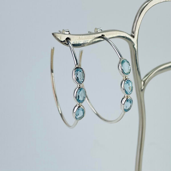 Large silver hoop earrings with three oval faceted blue topaz stones set into the front of the hoop. The stones are wider than the hoop and each is set into a frame of silver.