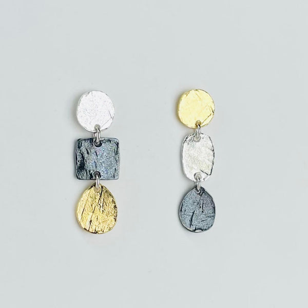 A pair of earrings that aren't identical! One has a bright  silver circle above a dark silver square above a gold plated pear shape. The other has a gold plated circle above a bright silver oval, above a dark silver pear shape. All are textured and joined by small jump rings. 