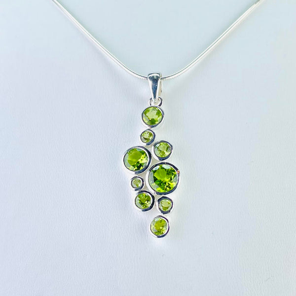 Lime green peridot pendant consisting of nine circular stones, all faceted and set in their own frames of silver. There are five different sizes of stone , 3 little, 3 small and three others each a little bigger than the last. They are randomly displayed down the pendant, the two bigger ones towards the centre with medium top and bottom and the smaller ones dotted in to make a vague diamond shape.