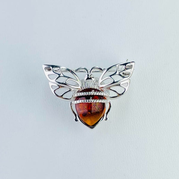 A silver and amber brooch modeled on a bee.  The body is pretty orange amber with two silver bands across the top and silver legs on either side. The wings  are outstretched,  cut out silver  and there is a little head and antennae at the top running parallel to the body.