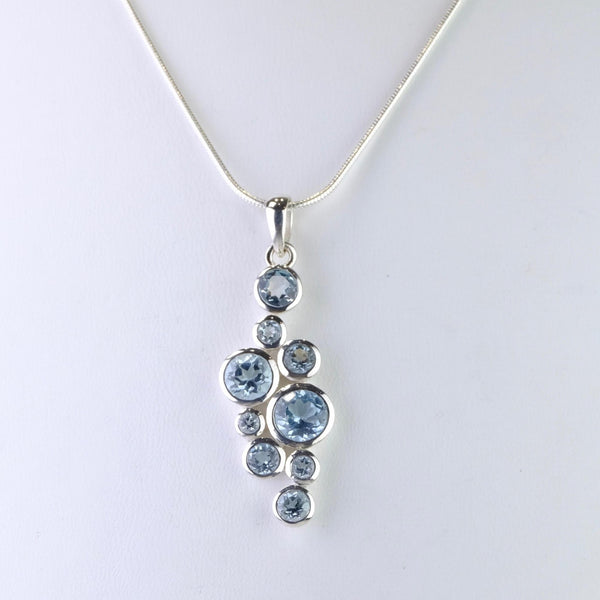 Blue topaz pendant consisting of nine circular stones, all faceted and set in their own frames of silver. There are five different sizes of stone , 3 little, 3 small and three others each a little bigger than the last. They are randomly displayed down the pendant, the two bigger ones towards the centre with medium top and bottom and the smaller ones dotted in to make a vague diamond shape.
