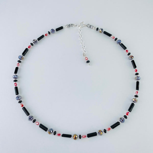 A necklace in black, grey and pink.  The pattern around the whole necklace is a fine glossy back tube bead, then a tiny silver ball, a small black circle. a tiny silver bead, a smokey grey faceted crystal, a tiny silver ball, a small deep pink faceted bead and a tiny silver ball. This pattern is repeated 14 times. It ends with a 2 inch silver link chain and  pink and grey beads .